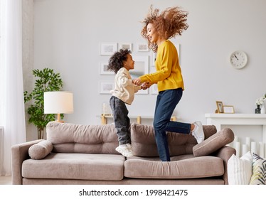 Crazy Weekend With Mom. Joyful Afro American Family Mother And Little Son Holding Hands And Jumping On Sofa In Living Room At Home, Full Length Shot. Mom And Child In Casual Wear Playing Together