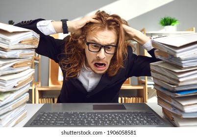 Crazy weird stressed young guy in glasses having busy day at work, sitting at office desk with computer, tearing hair in despair exhausted by much paperwork, huge file archive, red tape workload chaos