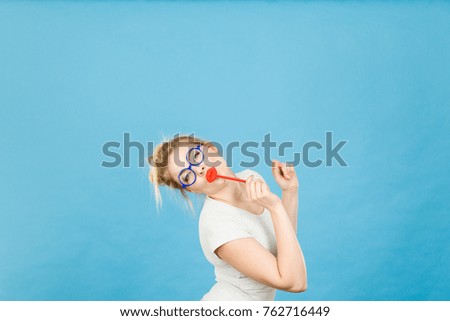Crazy woman casual style nerdy glasses holding red fake lips on stick having fun, on blue. Photo take and carnival funny accessories concept.