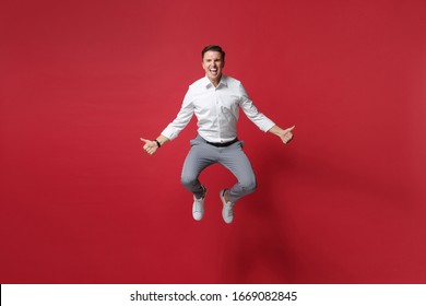 Crazy Young Business Man In White Shirt, Gray Pants Posing Isolated On Bright Red Background Studio Portrait. Achievement Career Wealth Business Concept. Mock Up Copy Space. Jumping Showing Thumbs Up