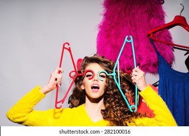 Crazy Young Girl With Curly Hair In Yellow Sweater Holding Hangers Standing Amid Colorful Clothes Pink Red Blue Colors On Grey Wall Background, Horizontal Picture