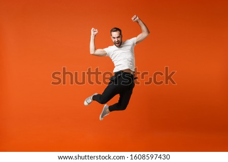 Crazy young man in casual white t-shirt posing isolated on orange background in studio. People lifestyle concept. Mock up copy space. Having fun, fooling around, jumping, clenching fists like winner
