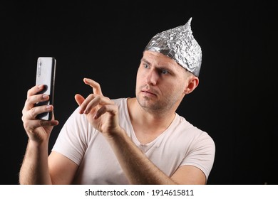 CrazyMan In A Foil Hat Holds Smartphone On Black Background. 5g. Conspiracy Theory