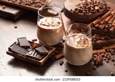 Cream and coffee cocktail in glasses with ice on an old wooden background. Coffee beans, cinnamon, anise, and pieces of chocolate are scattered on the table. 
