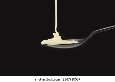 Cream Or Condensed Milk Pouring Down In A Black Spoon Against A Black Background.