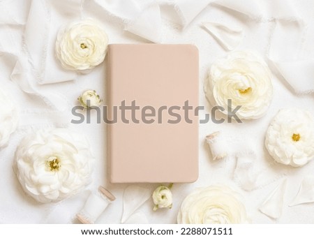 Cream hardcover textbook near cream roses and white silk ribbons top view,  mockup. Romantic scene with notebook and pastel flowers. Valentines, Spring or Mothers day concept