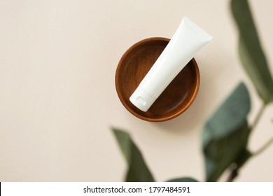 Cream Moisturizer On Wooden Plate On Light Beige Background With Blur Eucalyptus Leaf, Above. Natural Organic Eco Cosmetic Beauty Product. White Plastic Tube Mockup For Lotion, Cleanser Or Shampoo 