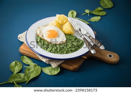 Creamed spinach with boiled potatoes and fried egg on email plate with cutlery, wooden board, napkin, raw spinach leaves and dark blue background