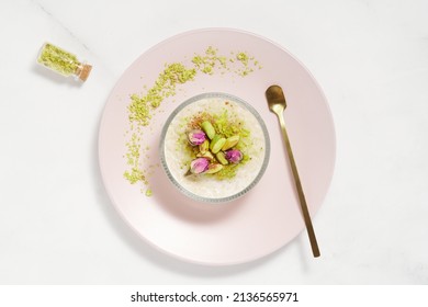 Creamy rice pudding topped with pistachio and cinnamon in a glass bowl on pink plate and marble table. Traditional Turkish dessert sutlac. Minimal bright and airy background.  Copy space. Top view.