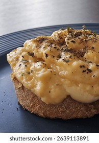 Creamy scrambled eggs served on toasted sourdough bread, topped with cracked black pepper and herbs. Simple and delicious breakfast dish, plated on a dark ceramic plate in natural light.