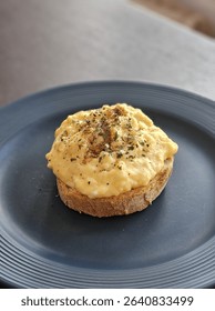 Creamy scrambled eggs served on toasted sourdough bread, topped with cracked black pepper and herbs. Simple and delicious breakfast dish, plated on a dark ceramic plate in natural light.