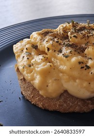 Creamy scrambled eggs served on toasted sourdough bread, topped with cracked black pepper and herbs. Simple and delicious breakfast dish, plated on a dark ceramic plate in natural light.