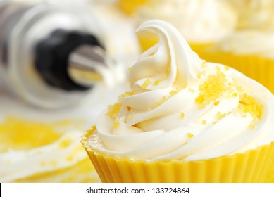 Creamy Swirl Of Vanilla Frosting On Cupcake In Silicone Baking Cup With Natural Lemon Sprinkles.  Cake Decorating Supplies In Soft Focus In Background.  Macro With Extremely Shallow Dof.