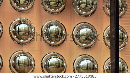 A creative abstract composition featuring a grid-like pattern of round, spherical convex mirrors mounted on a wall. Each mirror reflects a distorted fisheye view of modern urban architecture and a courtyard, creating a repetitive and visually striking geometric design with metallic silver accents.