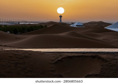 Creative alignment of a huge glowing sun resting perfectly on top of a distant landmark tower over dark sand dunes at sunset.
