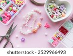 A creative arrangement of colorful beads, charms, and scissors on a pastel pink surface showcases the process of making unique jewelry. Delicate floral designs are highlighted.