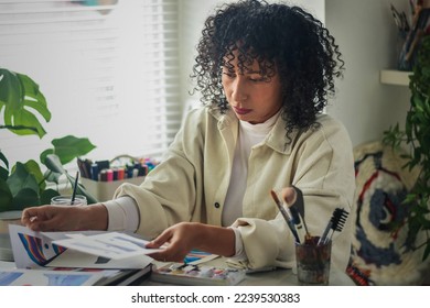 Creative artist diverse bi racial woman with curly hair in white room home studio by window sitting at desk surrounded by art tools, plants and shelf looking through art - Powered by Shutterstock