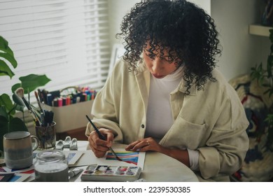 Creative artist diverse bi racial woman with curly hair in white room home studio by window sitting at desk surrounded by art tools, plants and shelf painting colourful lines on paper - Powered by Shutterstock