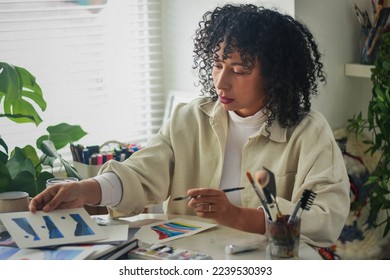 Creative artist diverse bi racial woman with curly hair in white room home studio by window sitting at desk surrounded by art tools, plants and shelf, picking select art work and painting - Powered by Shutterstock