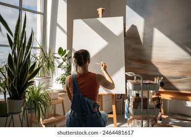 Creative artist woman starts to draw artwork on new white canvas on easel with brush at equipped modern art studio. Interested talented girl painter enjoy creative hobby for soul in painter workshop. - Powered by Shutterstock