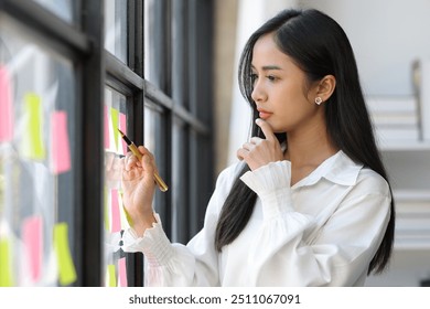 Creative Asian businesswoman thinking and planning startup business with paper stickers on glass, analyzing strategy and planning business in office. - Powered by Shutterstock