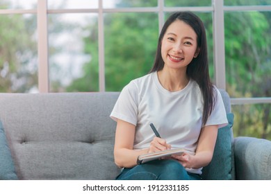 Creative Asian Woman Is Writing Memory Planning And Idea On To A Notebook On Sofa At Home.