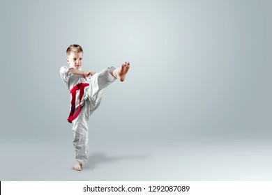 Creative Background, A Child In A White Kimono Makes A Kick, On A Light Background. The Concept Of Martial Arts, Karate, Sports Since Childhood, Discipline, First Place, Victory. Copy Space.