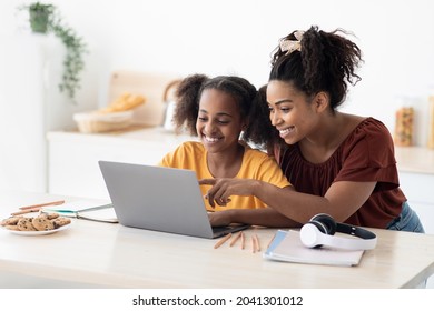Creative black mother and daughter working on school project, using modern laptop, taking notes, cheerful afro-american woman helping her teen kid with homework, kitchen table, copy space