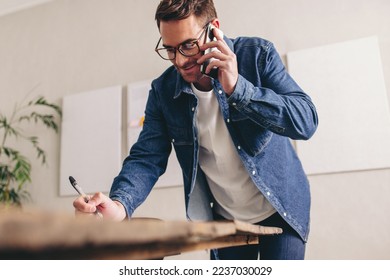 Creative businessman writing notes during a phone call in his office. Happy young businessman making plans with his clients over the phone. Young businessman with glasses working remotely. - Powered by Shutterstock