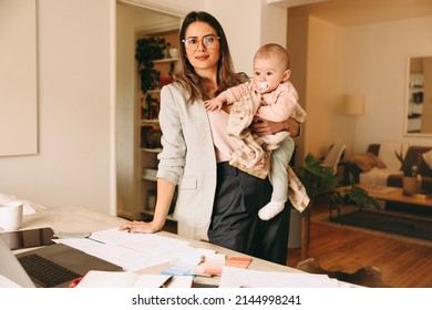 Creative businesswoman holding her baby while standing behind her desk. Working mom planning a new project in her home office. Female interior designer balancing work and motherhood.