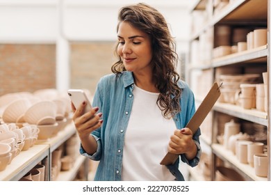 Creative businesswoman using a smartphone in her ceramic store. Cheerful entrepreneur managing a shop with handmade ceramic products. Young female ceramist running a successful small business.