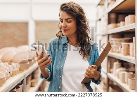 Creative businesswoman using a smartphone in her ceramic store. Cheerful entrepreneur managing a shop with handmade ceramic products. Young female ceramist running a successful small business.