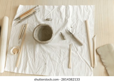 Creative clay pot on worktable with wooden handles and cloth, top-down perspective, gray and white tones, artistic and serene mood, crafting and pottery context.