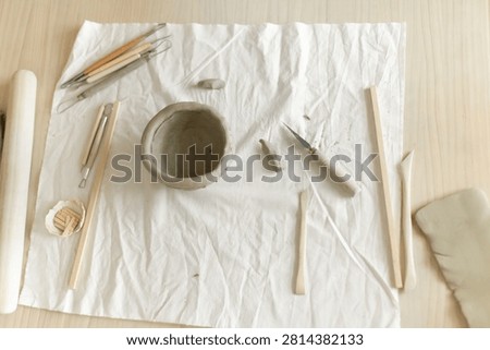 Creative clay pot on worktable with wooden handles and cloth, top-down perspective, gray and white tones, artistic and serene mood, crafting and pottery context.