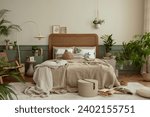 Creative composition of bedroom interior with cozy bed, plants, beige beding, stylish lamp, rattan armchair, pouf, carpet, brown slippers, wooden stool and personal accessories. Home decor. Template.