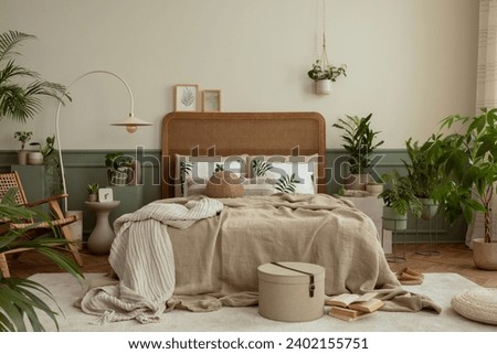 Creative composition of bedroom interior with cozy bed, plants, beige beding, stylish lamp, rattan armchair, pouf, carpet, brown slippers, wooden stool and personal accessories. Home decor. Template.