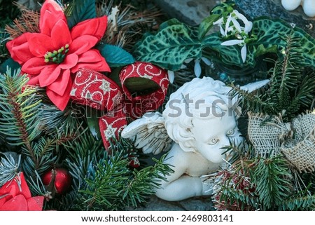 Creative composition with cute white angel laying on a gravestone, surrounded with pine branches and red Poinsettia flowers, red ribbon, Christmas decor. 