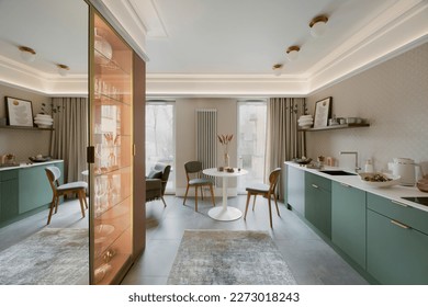 Creative composition of dinning room interior with kitchen space, round table, chair, green shelf, vase with dried flowers, gray floor, rug, glass shelf and personal accessories. Home decor. Template.