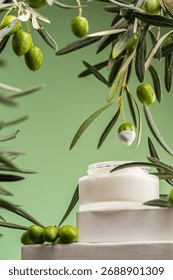 Creative composition with a fresh olive dipped in white cosmetic cream above an open skincare jar surrounded by olive branches on a green background. Concept of natural beauty products,
