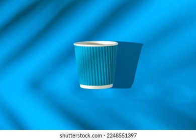 Creative composition made of paper cup of coffee or tea to go on bright blue background with sunlit and palm shadow. Eco friendly concept. Minimal style. Flat lay. Top view