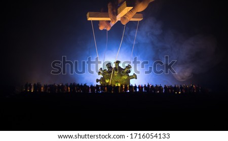 Creative concept of manipulation with coronavirus. Crowd looking on big Corona virus miniature. The hand controls the Covid19 miniature with strings on a dark foggy background. Selective focus