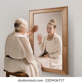 Creative conceptual collage. Tender image of senior woman looking in mirror and smiling to her young self reflection. Back to past. Concept of present, past and future, age, lifestyle, memories, ad - Powered by Shutterstock