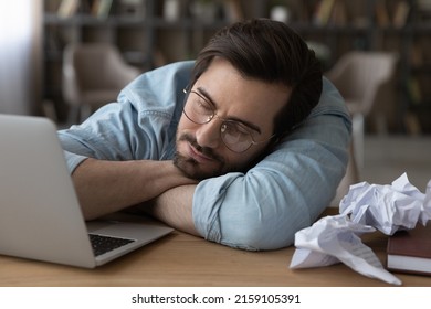 Creative Crisis. Tired Young Male Writer Freelancer Lie On Work Desk Sleep By Laptop Among Crumpled Papers Feel Mental Block. Frustrated Employee Fall Asleep At Workplace Having Professional Burnout