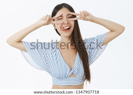 Creative delighted and chill asian woman in stylish cropped top sticking out tongue joyfully winking showing peace or victory signs over eyes dancing disco and having fun enjoying life to fullest