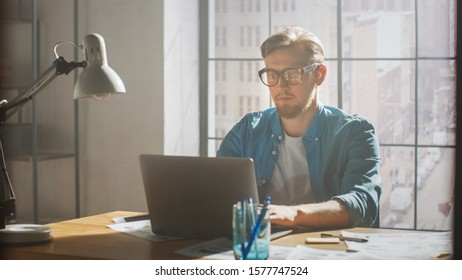 Creative Developer Works On A Laptop In His Studio. Designing Apps, Games And Doing Video Editing. In The Background Modern Sunny Loft Office With Project Storyboard And Concepts Covering The Wall