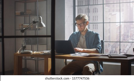 Creative Developer Works On A Laptop In His Studio. Designing Apps, Games And Doing Video Editing. In The Background Modern Sunny Loft Office With Project Storyboard And Concepts Covering The Wall