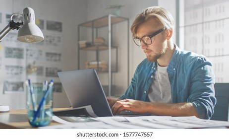 Creative Developer Works On A Laptop In His Studio. Designing Apps, Games And Doing Video Editing. In The Background Modern Sunny Loft Office With Project Storyboard And Concepts Covering The Wall