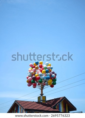 A creative display featuring an artificial bouquet of colorful balloons attached to the roof of a small replica hut. The scene combines playful imagination with rustic charm, highlighting a unique art