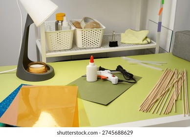 A creative DIY project in progress. A vibrant green desk holds a hot glue gun, glue sticks, colored paper, and wooden dowels. Bright, focused lighting from a desk lamp illuminates the workspace.