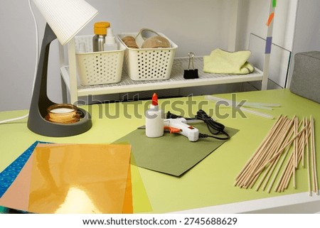 A creative DIY project in progress. A vibrant green desk holds a hot glue gun, glue sticks, colored paper, and wooden dowels. Bright, focused lighting from a desk lamp illuminates the workspace.